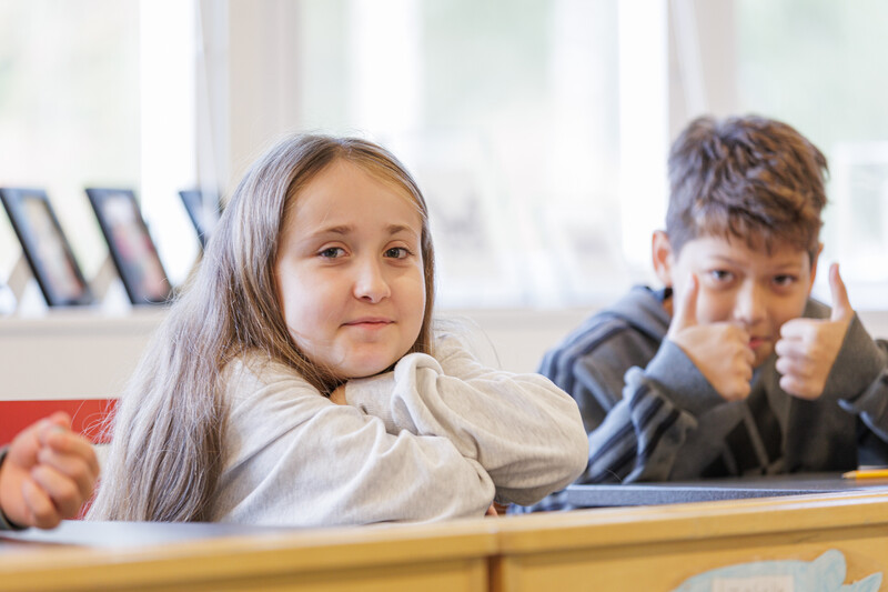 En elev med långt hår tittar leende in i kameran, medan en klasskamrat bredvid gör tumme upp, båda sittandes vid sina bänkar i ett ljust klassrum.