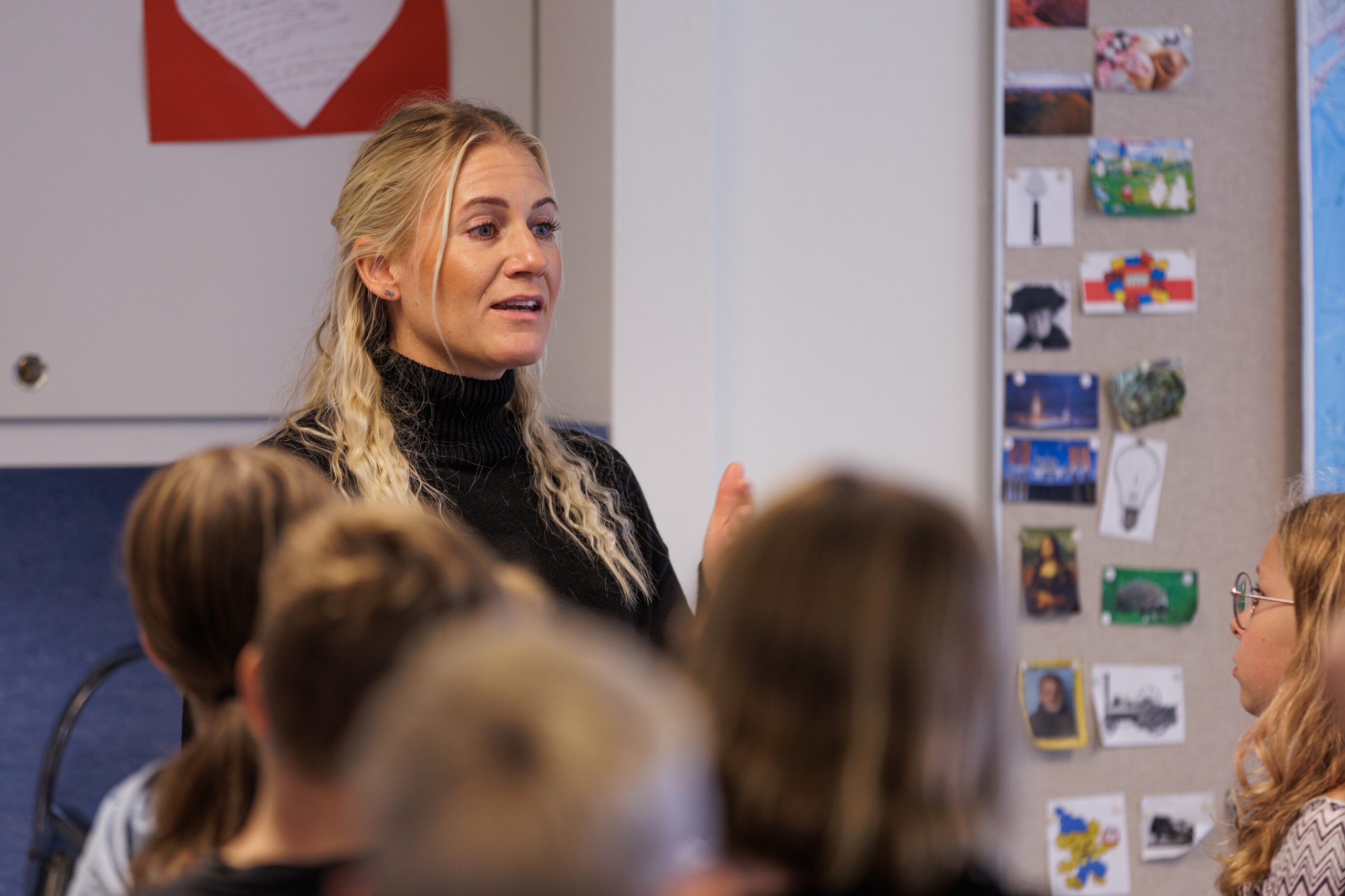 En lärare med blont hår pratar och gestikulerar framför en grupp elever i ett klassrum, med teckningar och bilder uppsatta på en anslagstavla i bakgrunden.