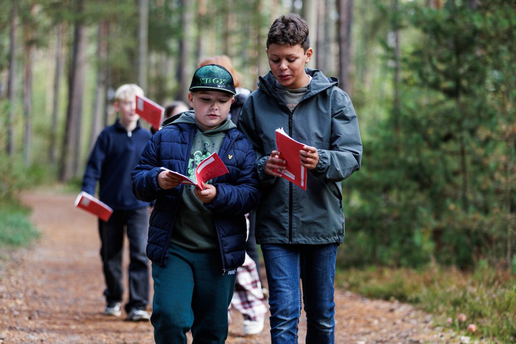 Två barn går tillsammans på en skogsstig och läser i röda häften, med ytterligare ett barn synligt längre bak på stigen bland träden.