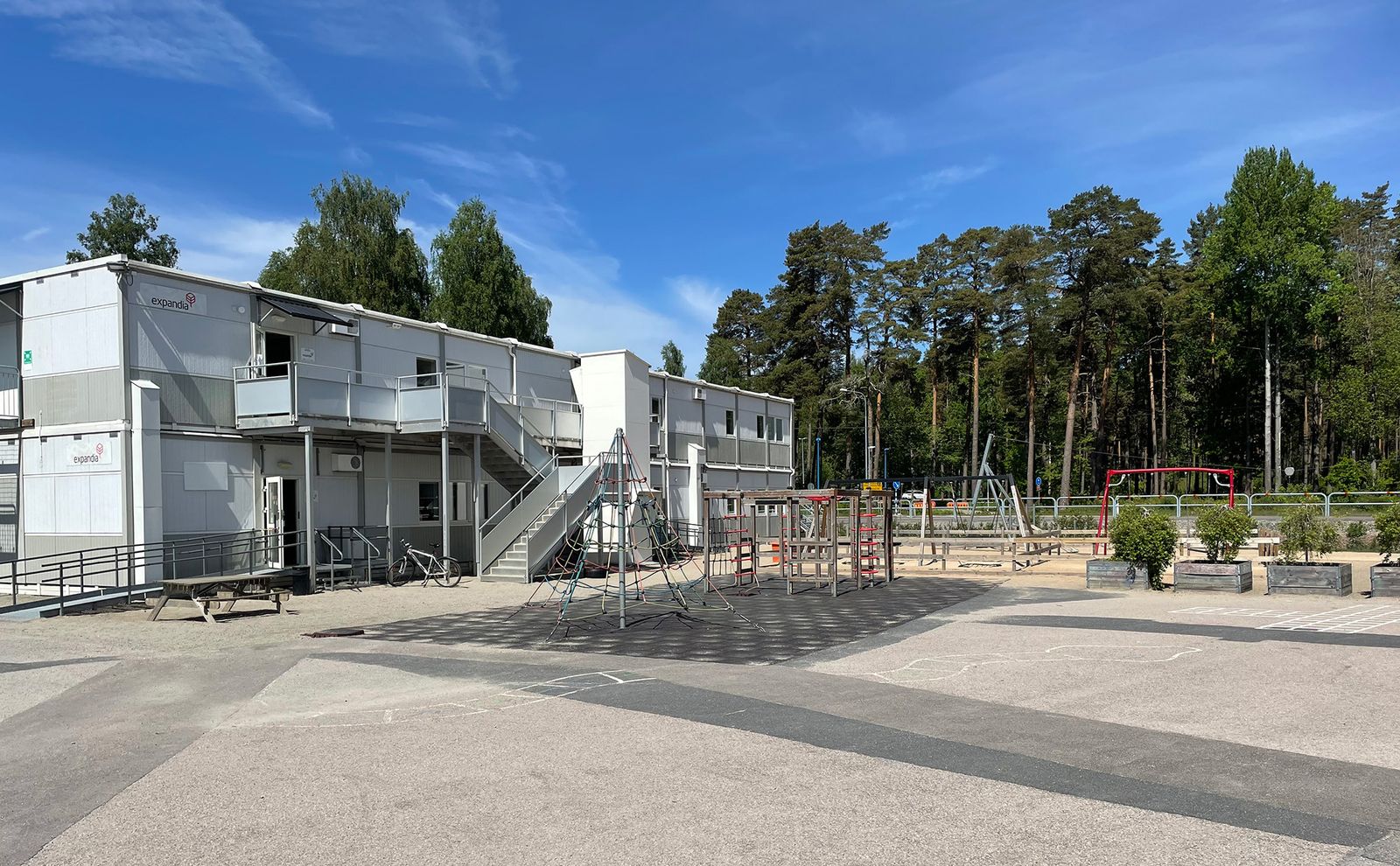 Hagatorpskolans skolbyggnad i modulform med utomhustrappa, med en lekplats med klätterställningar och gungor på skolgården samt en tallskog i bakgrunden en klarblå dag.